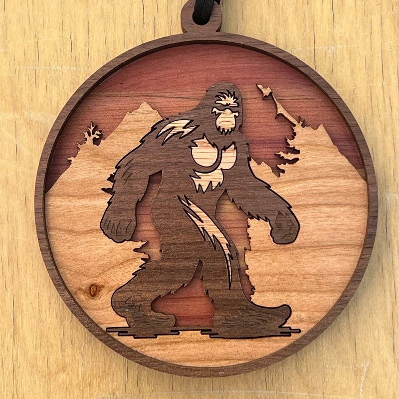 A round wooden pendant ornament featuring a laser-cut silhouette of a Bigfoot figure against a mountain landscape, hanging from a black cord, shown on a light wood background.