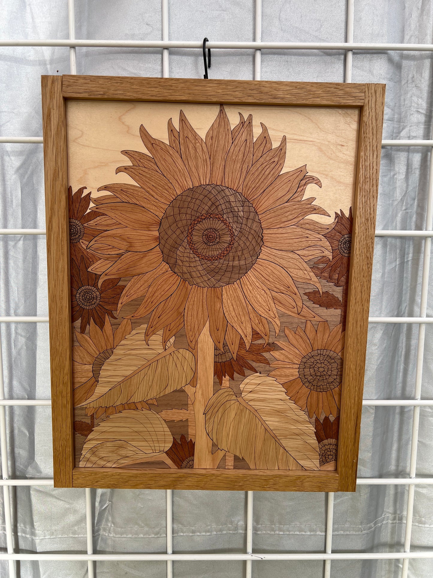 A rectangular wooden wall art piece featuring an intricate sunflower and leaf inlay design, mounted on a white wire grid background.
