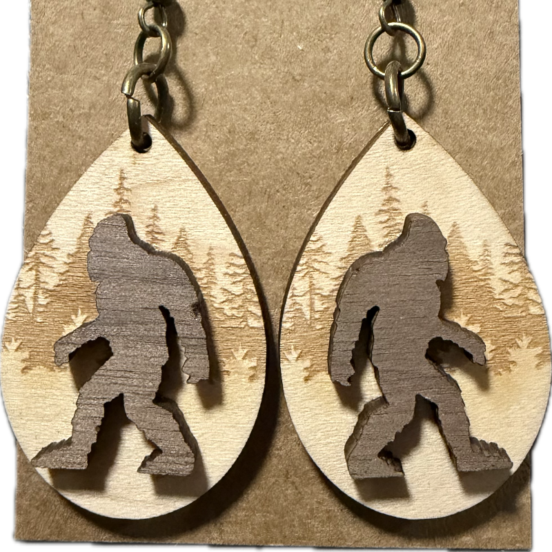 A pair of teardrop-shaped wooden dangle earrings displayed on a kraft paper backing card. The earrings are made from light natural wood with a darker brown laser-cut silhouette of Bigfoot and a forest pattern in the background, attached to antique bronze fishhook ear wires.