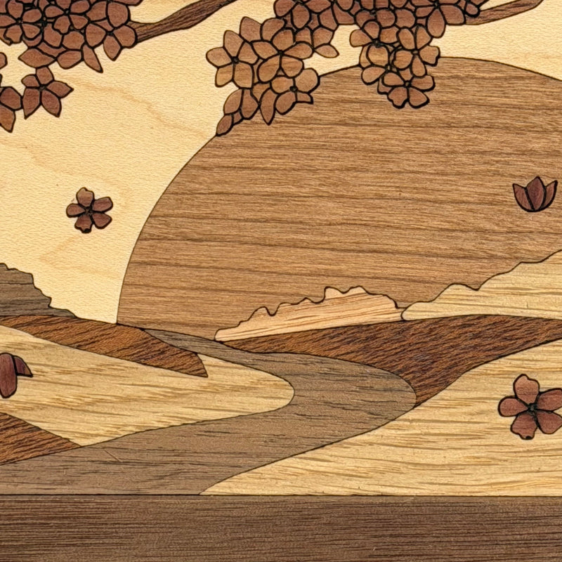 Close-up of an inlaid wooden landscape artwork with cherry blossom branches, a large sun or moon shape, rolling hills, and a winding stream in varying wood tones.