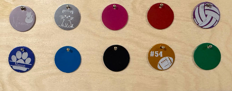 A set of round metal pet ID tags with hanging loops, arranged in two rows on a light wood background. The tags feature various colors and designs, including solid pink, red, blue, black, green, purple-and-white volleyball pattern, paw prints, and cat and cat-ear illustrations.