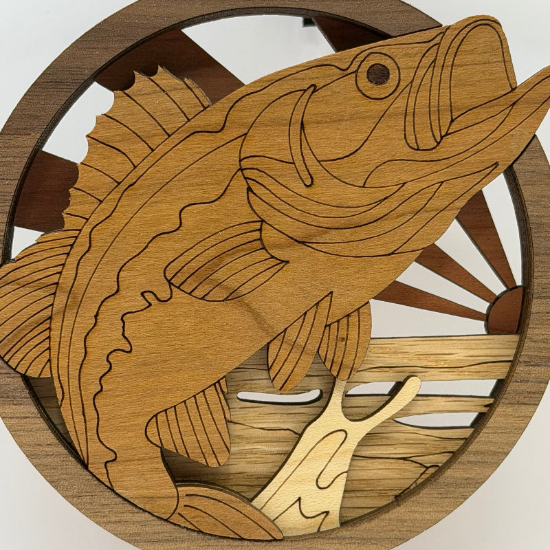 A circular wooden wall art piece featuring a laser-cut bass fish in natural wood tones, with a sunburst and water design in the background.