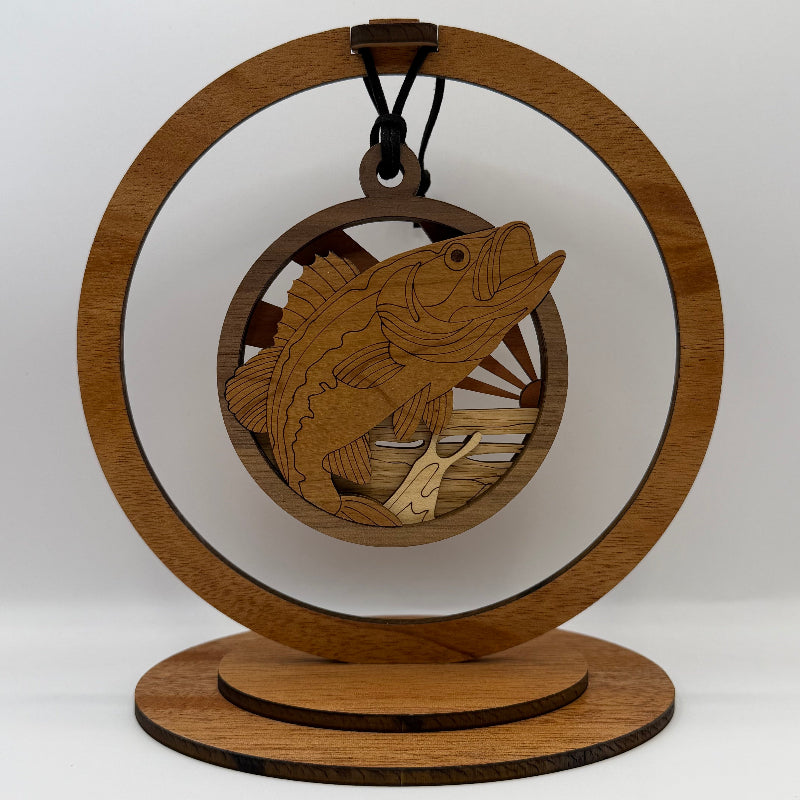 A laser-engraved wooden ornament shaped like a fish jumping from water, hanging from a black cord inside a circular wooden display stand with a round base, photographed against a light gray studio background.