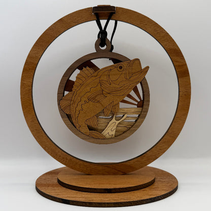A laser-engraved wooden ornament shaped like a fish jumping from water, hanging from a black cord inside a circular wooden display stand with a round base, photographed against a light gray studio background.