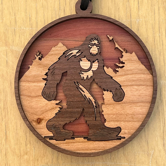A round wooden pendant ornament featuring a laser-cut silhouette of a Bigfoot figure against a mountain landscape, hanging from a black cord, shown on a light wood background.