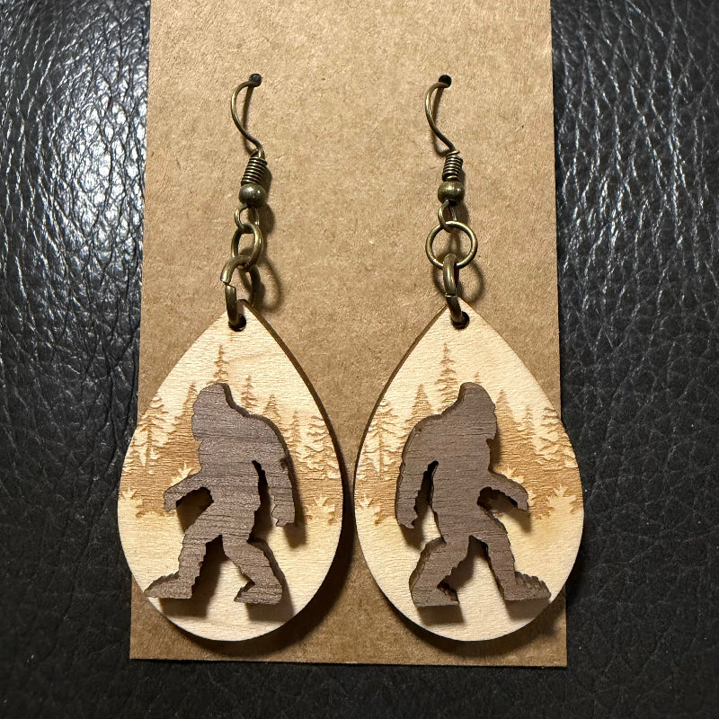 A pair of dangling teardrop earrings displayed on a kraft paper card against a black textured background. The earrings are made from light natural wood with a darker brown silhouette of Bigfoot cut out in the center, and hang from dark metal fishhook wires.