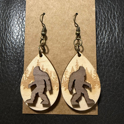 A pair of dangling teardrop earrings displayed on a kraft paper card against a black textured background. The earrings are made from light natural wood with a darker brown silhouette of Bigfoot cut out in the center, and hang from dark metal fishhook wires.