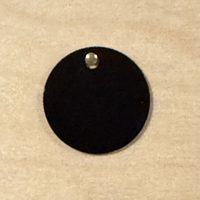 A black round metal tag with a small hole at the top, shown on a light wood background.