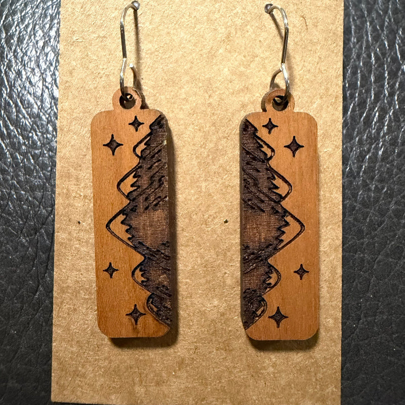 A pair of wooden rectangular drop earrings with dark brown engraved mountain and star designs, hanging from silver fishhook wires on a brown kraft paper display card.