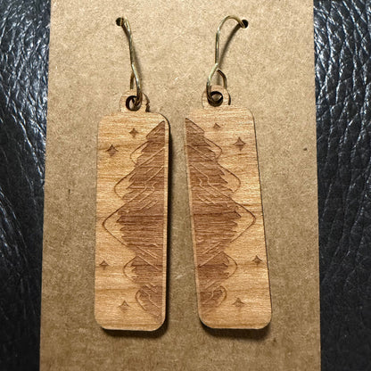 A pair of rectangular wooden drop earrings with engraved geometric and tree-like patterns, hanging from metal hooks on a brown kraft paper display card against a black textured background.