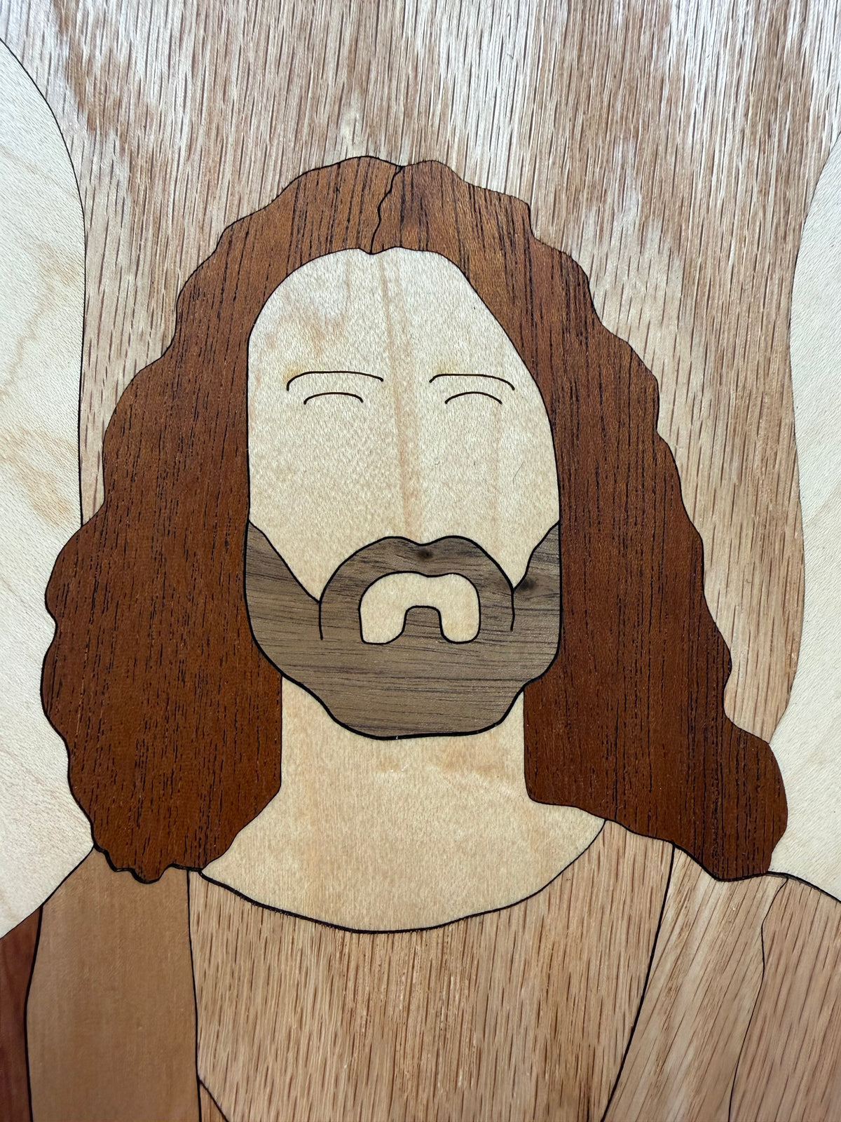 A close-up of a wooden portrait artwork showing a face with long wavy hair, a beard, and a simple garment, crafted from different wood tones.
