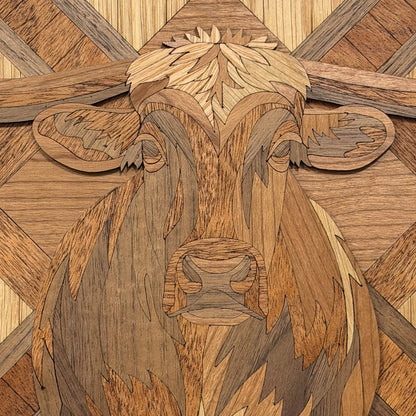 A close-up of a wooden cow face artwork made from layered wood in multiple warm tones, with carved details for the eyes, ears, nose, and textured fur, set against a geometric wood pattern background.