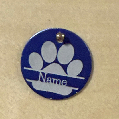 A round blue pet ID tag with a white paw print and the customizable text 'Name' in the center, attached to a small metal loop at the top, shown on a beige background.