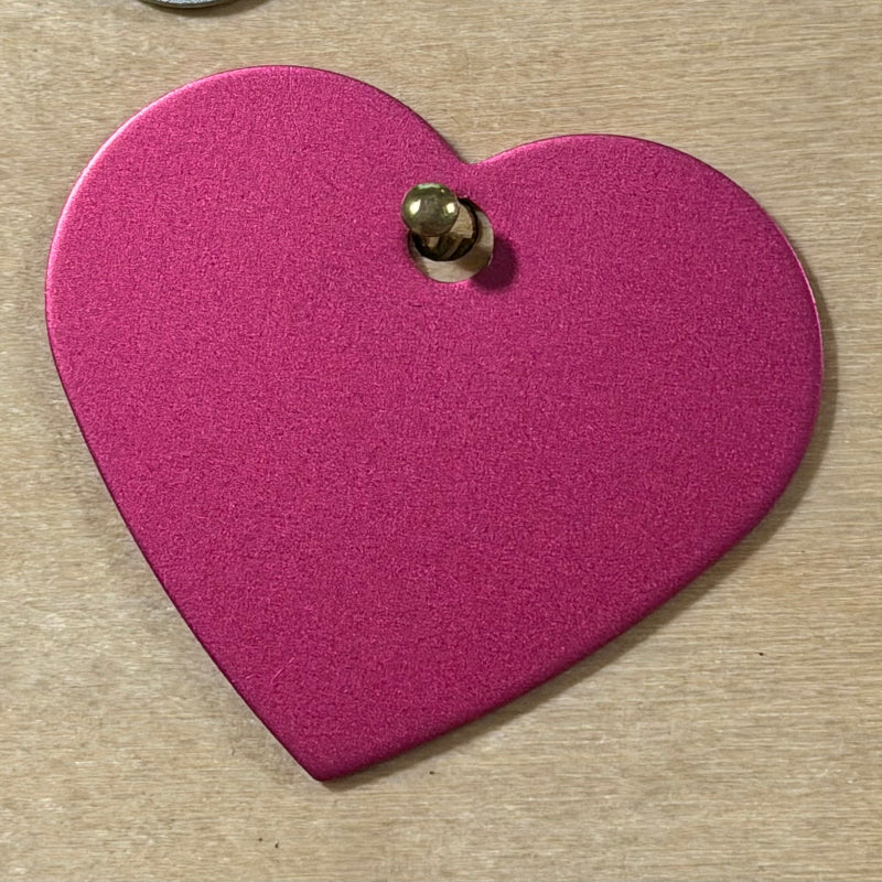 A dark pink heart-shaped metal tag with a small hole at the top, shown on a light wood background.