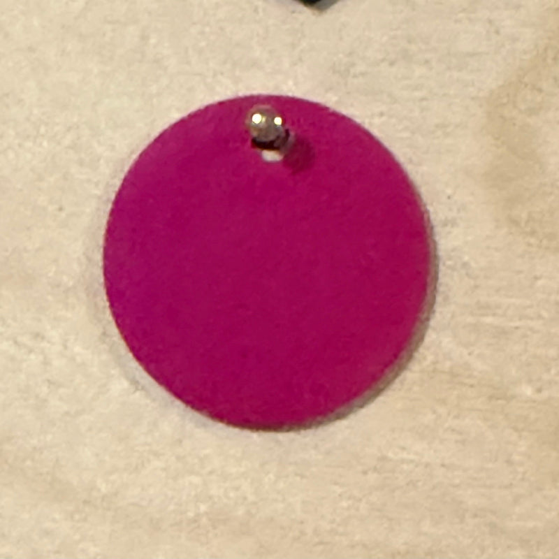 A dark pink round metal pet tag with a small hole at the top, shown on a light wood background.