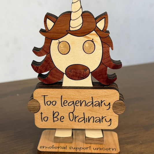 A wooden unicorn figurine with a scroll that reads 'Too legendary to Be Ordinary' and a base that says 'emotional support unicorn', displayed on a wooden surface against a light wall.