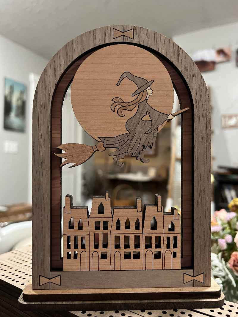 Wooden, multi-layerd decorative arch that sits on a shelf, desk, or table with a  witch in front of a full moon flying over a town