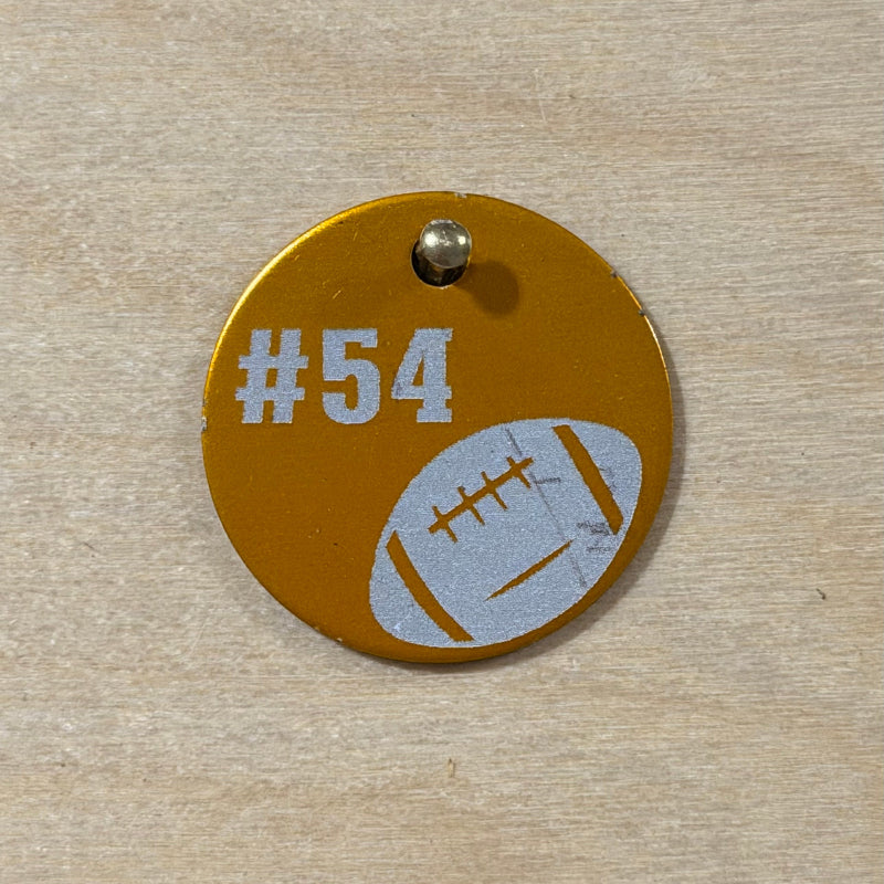 A round gold-colored metal pet ID tag with a silver screw, featuring the text '#54' and a white football graphic, shown on a light wood background.