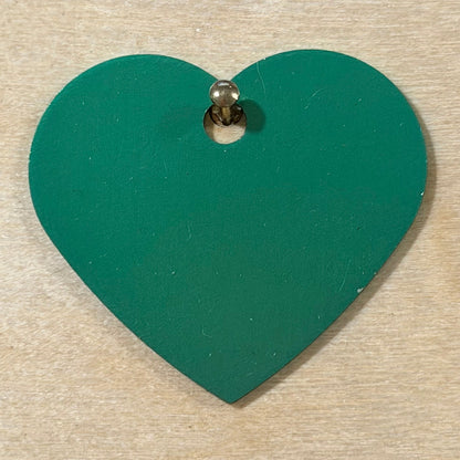 A green heart-shaped wooden tag with a pre-drilled hole, shown on a light wood grain background.