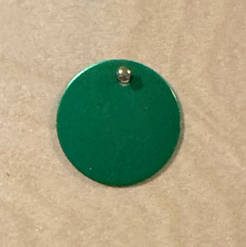 A green round metal pet tag with a small hole at the top, shown on a light wood background.