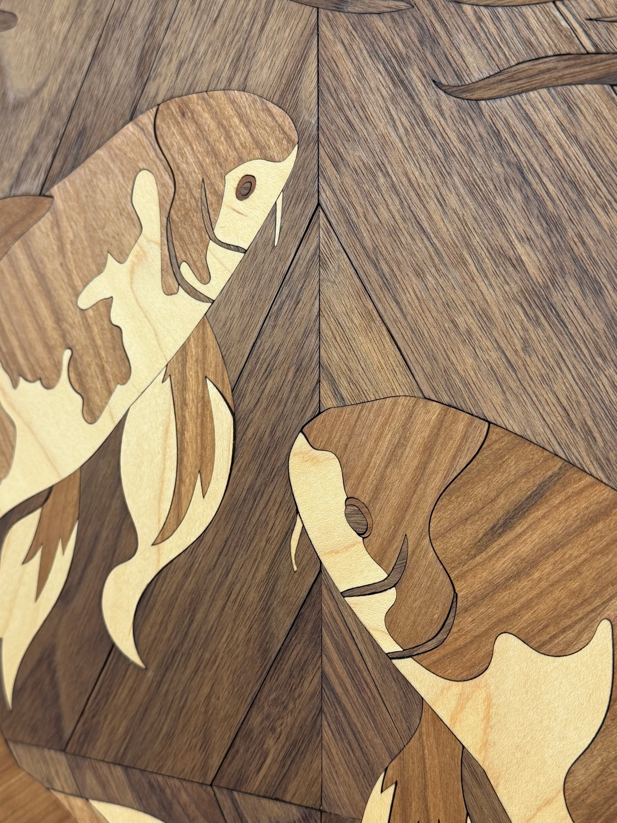 Close-up of a wooden inlay design featuring stylized koi fish in warm medium brown and pale cream tones against a darker wood grain background.