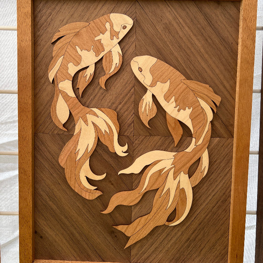 A handmade wooden wall art piece featuring two stylized koi fish inlaid into a dark wood background, photographed against a white tiled wall.