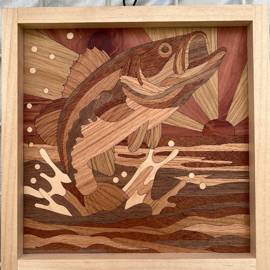 A square wooden framed handmade artwork featuring an inlaid bass fish and natural wood grain patterns, displayed against a light gray fabric background.
