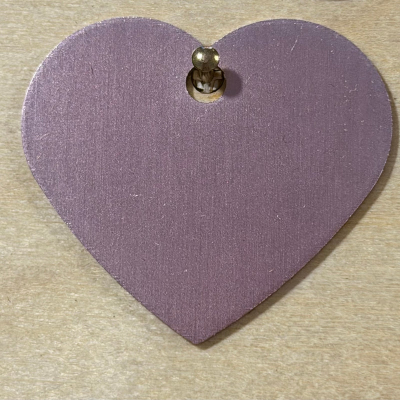 A light pink heart-shaped metal tag with a small hole at the top, shown on a light wood background.