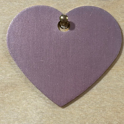 A light pink heart-shaped metal tag with a small hole at the top, shown on a light wood background.