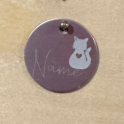 A round metal pet ID tag with a light pink background, featuring a white cat silhouette with a heart on its belly and the word 'Name' in cursive script, on a light wood background