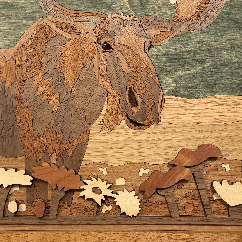 A close-up of a wooden moose head and antlers carved from layered wood tones, with autumn leaves and acorns arranged in front on a wooden surface.