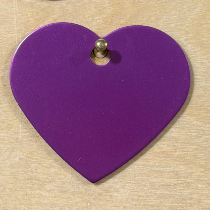 A heart-shaped purple metal tag with a pre-drilled hole, shown on a light wood background.