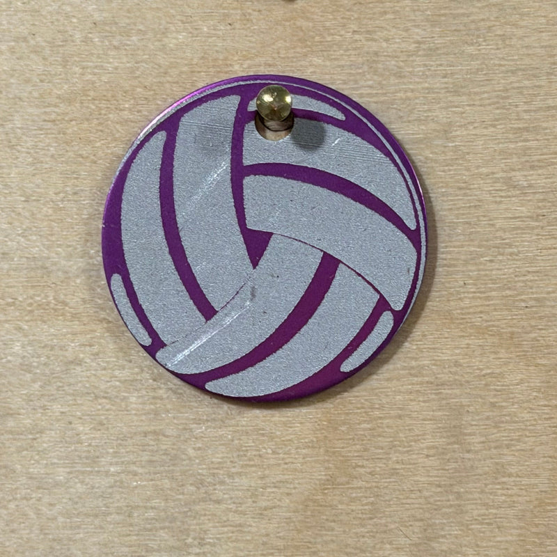 A round metal pet tag featuring a purple and silver volleyball pattern, photographed on a light wood grain surface.