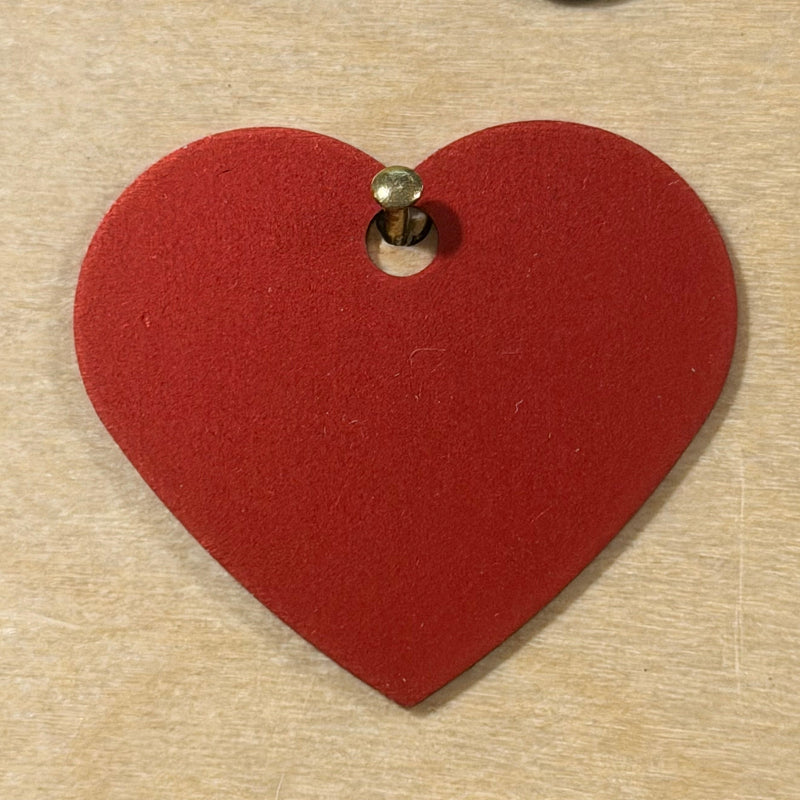 A red heart-shaped metal tag with a pre-drilled hole shown on a light wood background.