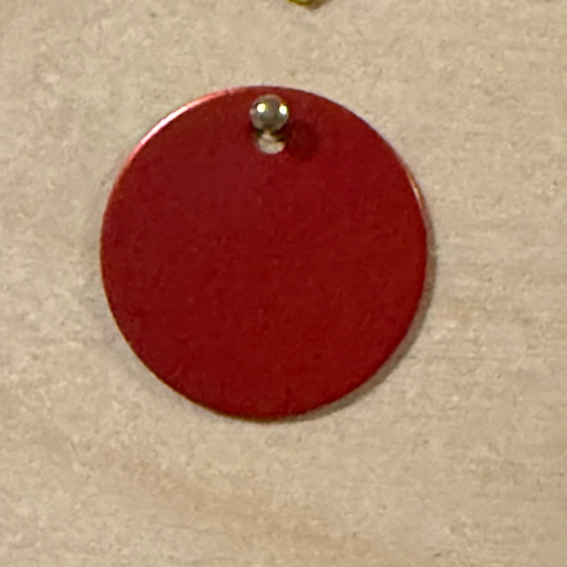 A red round metal pet tag with a small hole at the top, shown on a light wood background.