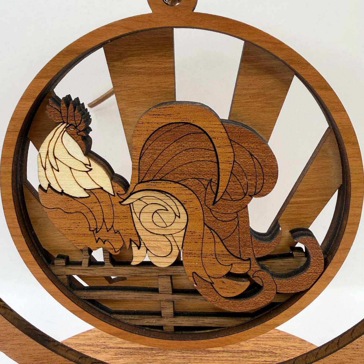 A circular wooden rooster figurine with layered laser-cut details, displayed on a wooden stand against a white background.