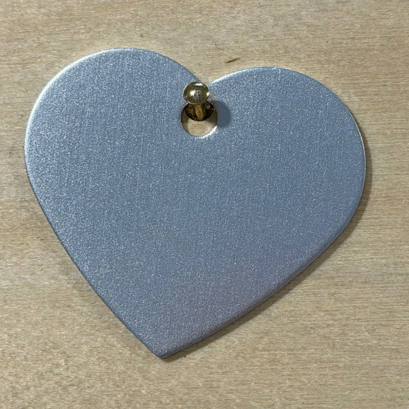 A heart-shaped metallic silver tag with a small gold-colored screw fastener at the top, shown on a light wood background.