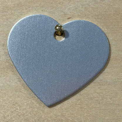 A heart-shaped metallic silver tag with a small gold-colored screw fastener at the top, shown on a light wood background.