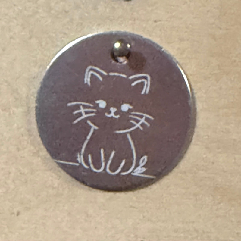 A round metal dog tag with a small cat outline engraved on it, featuring a top loop and photographed on a tan textured surface.