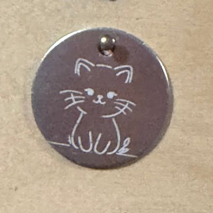 A round metal dog tag with a small cat outline engraved on it, featuring a top loop and photographed on a tan textured surface.
