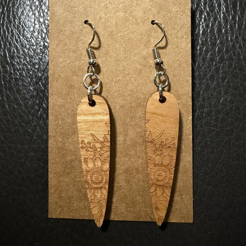 A pair of wooden dangle earrings with floral engravings, hanging from silver-toned fishhook wires on a brown kraft display card, set against a dark textured background.