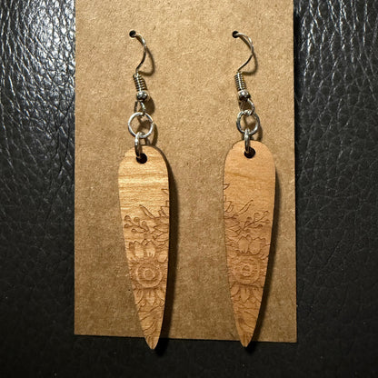 A pair of wooden dangle earrings with floral engravings, hanging from silver-toned fishhook wires on a brown kraft display card, set against a dark textured background.