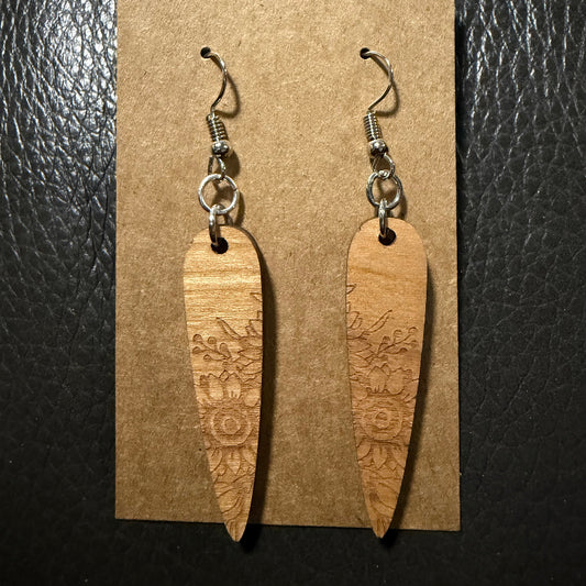 A pair of wooden dangle earrings with floral engravings, hanging from silver-toned fishhook wires on a brown kraft display card, set against a dark textured background.