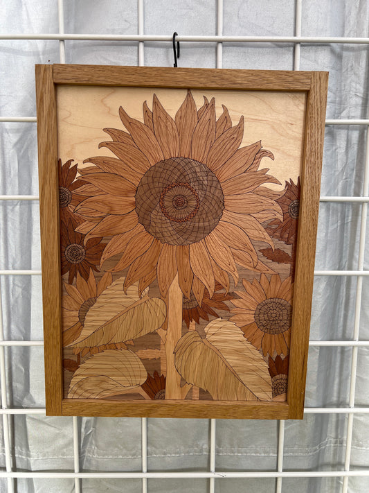 A rectangular wooden wall art piece featuring an intricate sunflower and leaf inlay design, mounted on a white wire grid background.
