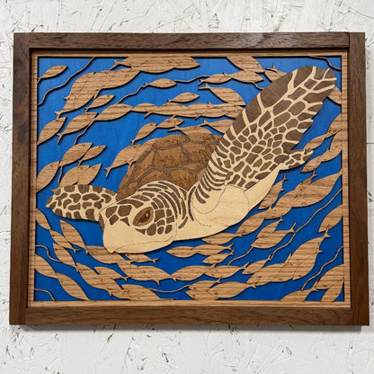 A square framed wooden wall art piece featuring a sea turtle layered over blue and tan wood cutouts, mounted on a white textured wall.
