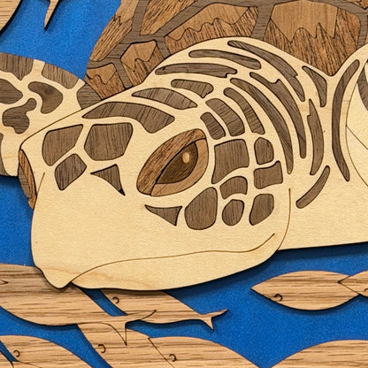 Close-up of a wooden sea turtle artwork with layered laser-cut wood pieces in natural tan and medium brown tones, set against a bright blue background with additional fish shapes.