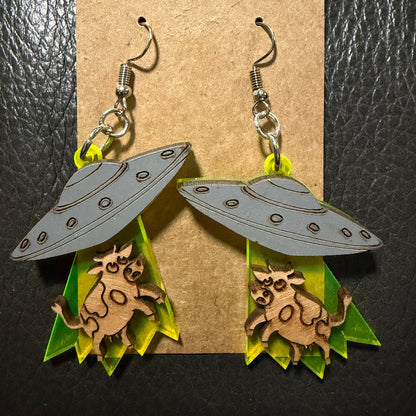 gray UFO lifting a cow in it's green acrylic beam  earrings on a craft paper card with a black textured backgournd