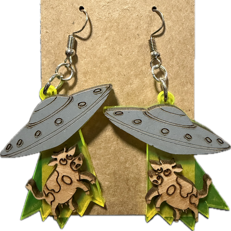 A pair of dangling earrings displayed on a kraft paper backing card. Each earring has a gray UFO-shaped top with small circular details, a wooden cow figure in the center, and layered neon yellow-green triangular shapes below.