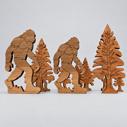 A wooden cutout scene featuring two big-footed figures walking among pine trees, displayed on a dark wooden surface against a light textured wall.