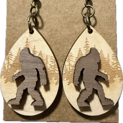 A pair of teardrop-shaped wooden dangle earrings displayed on a kraft paper backing card. The earrings are made from light natural wood with a darker brown laser-cut silhouette of Bigfoot and a forest pattern in the background, attached to antique bronze fishhook ear wires.